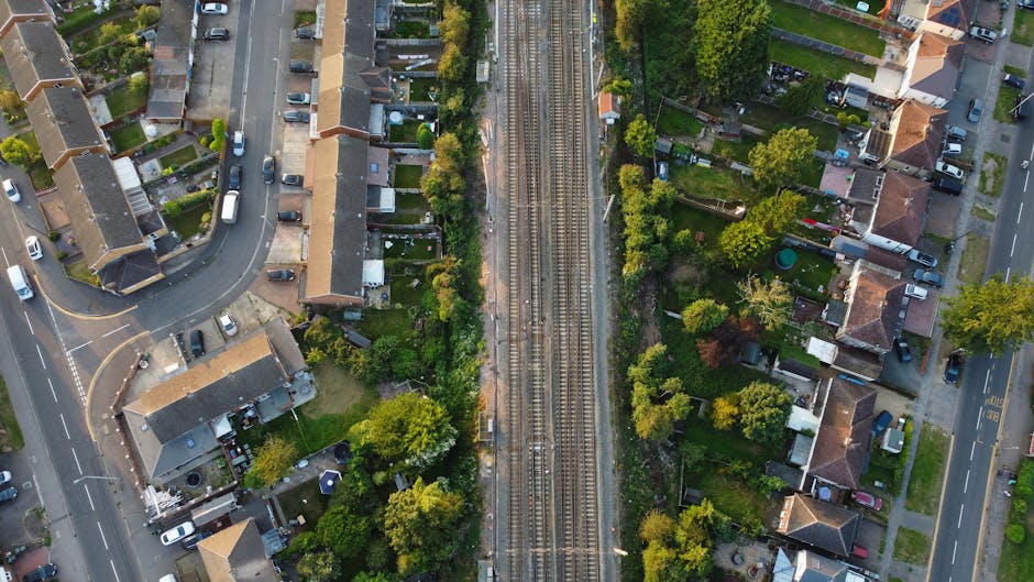 An aerial view of a suburban neighborhood showing a railway line running vertically through the center, flanked by residential houses with well-maintained gardens, trees, and lawns. The houses on the 